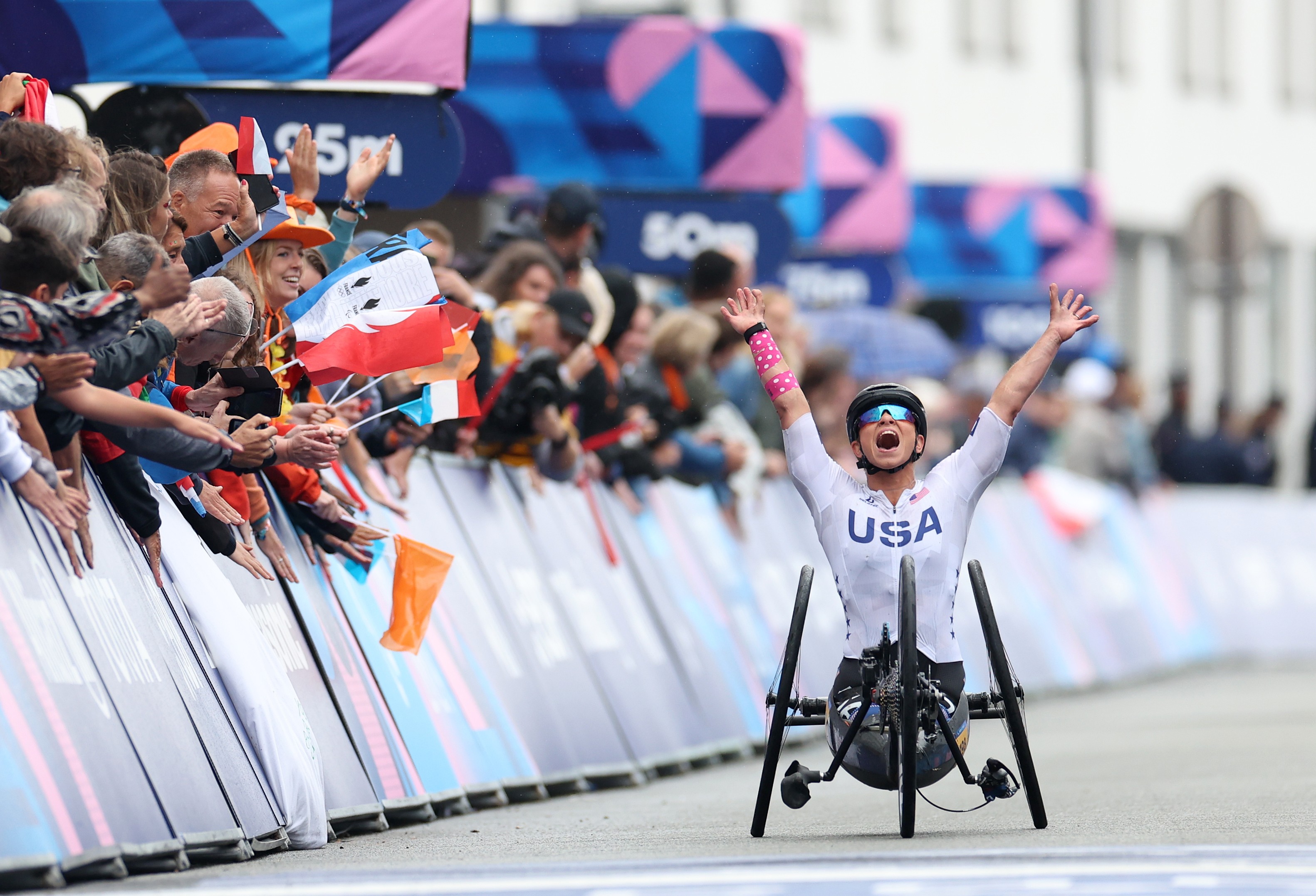 An athlete in a racing wheelchair raises their arms in celebration as they pass a crowd at a finish line.