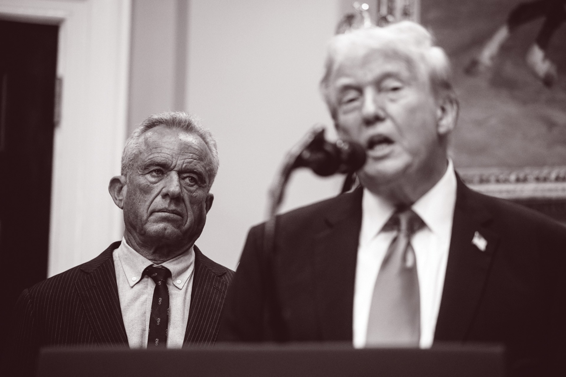 black-and-white photo of Trump speaking into microphone at podium with RFK Jr. looking on from background