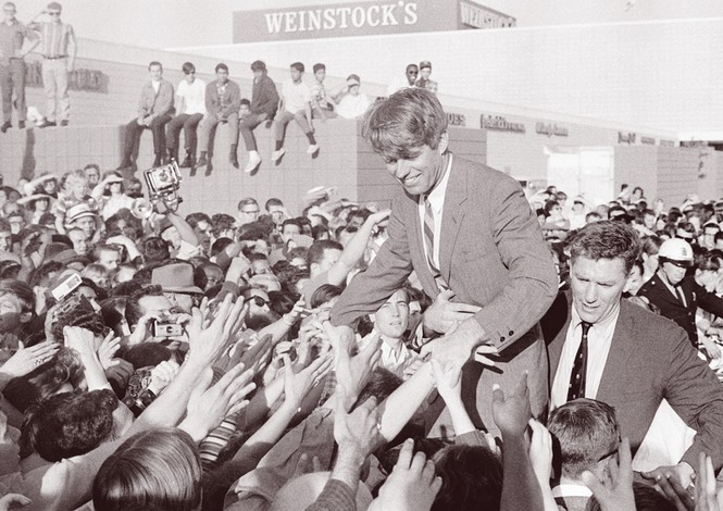 black-and-white archival photo of RFK Sr. smiling and standing above crowd of people with outstretched arms and shaking hands